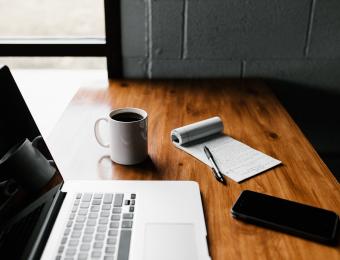 Image of a computer, coffee mug, pad of paper with notes on it, and phone on a desk